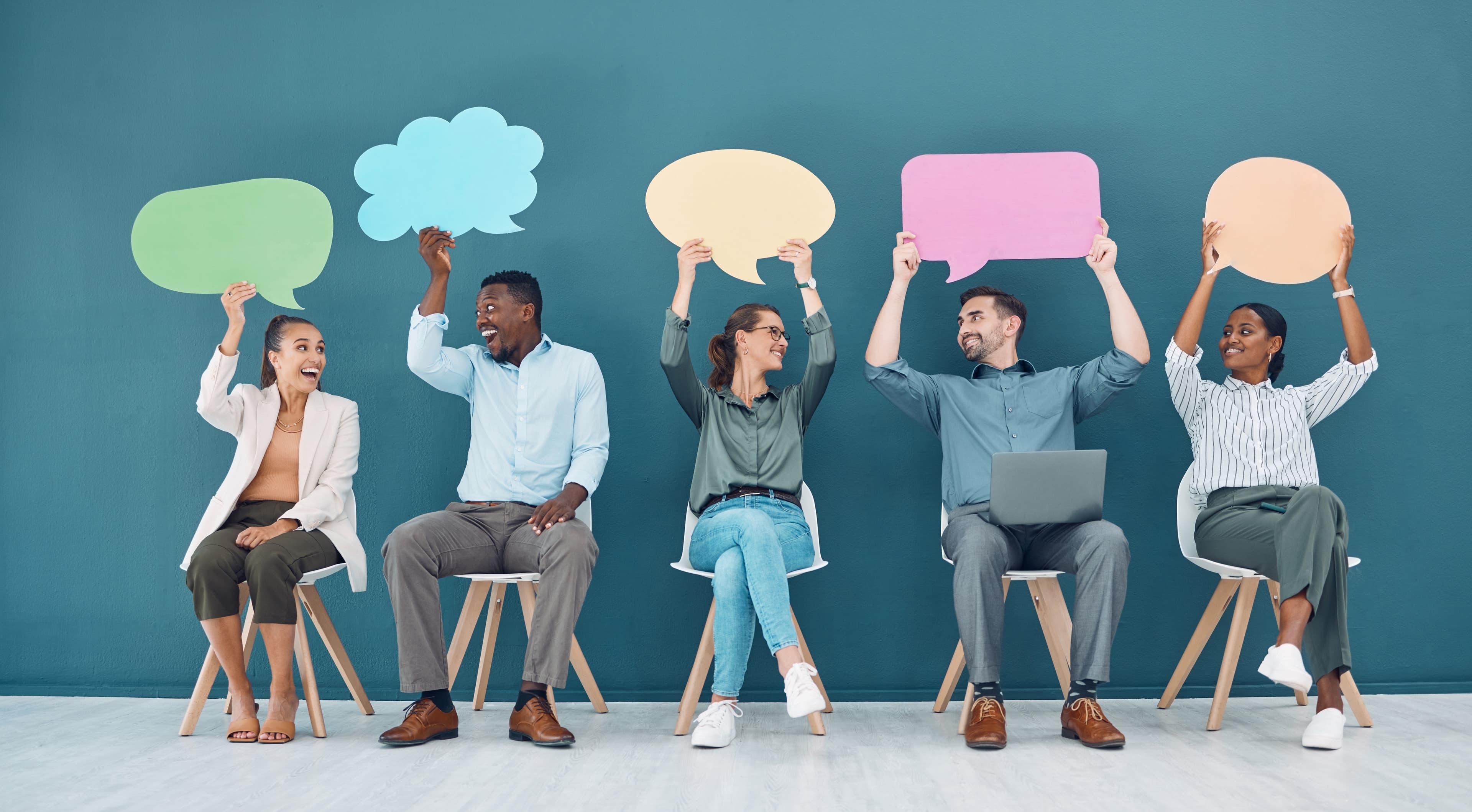 Five people seated on chairs holding colored speech bubbles above their heads against a teal wall.
